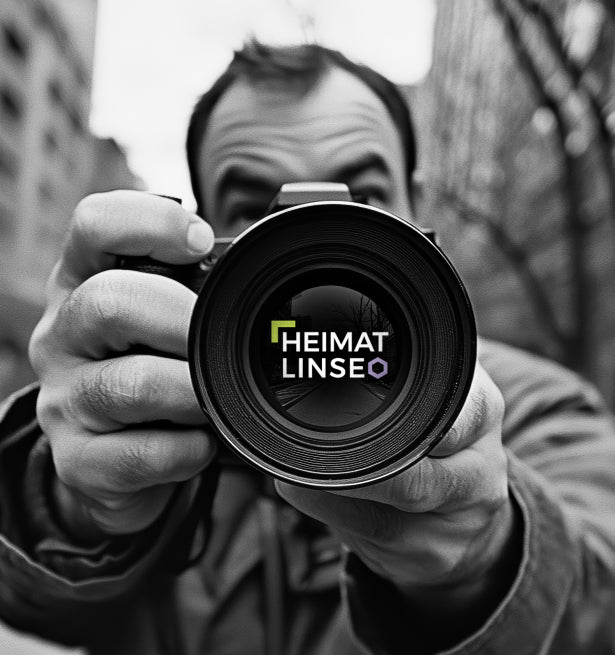 Graues Bild eines Mannes umgeben von Gebäuden, der eine Kamera hält, in dessen Linse das Heimatlinse-Logo abgebildet ist.