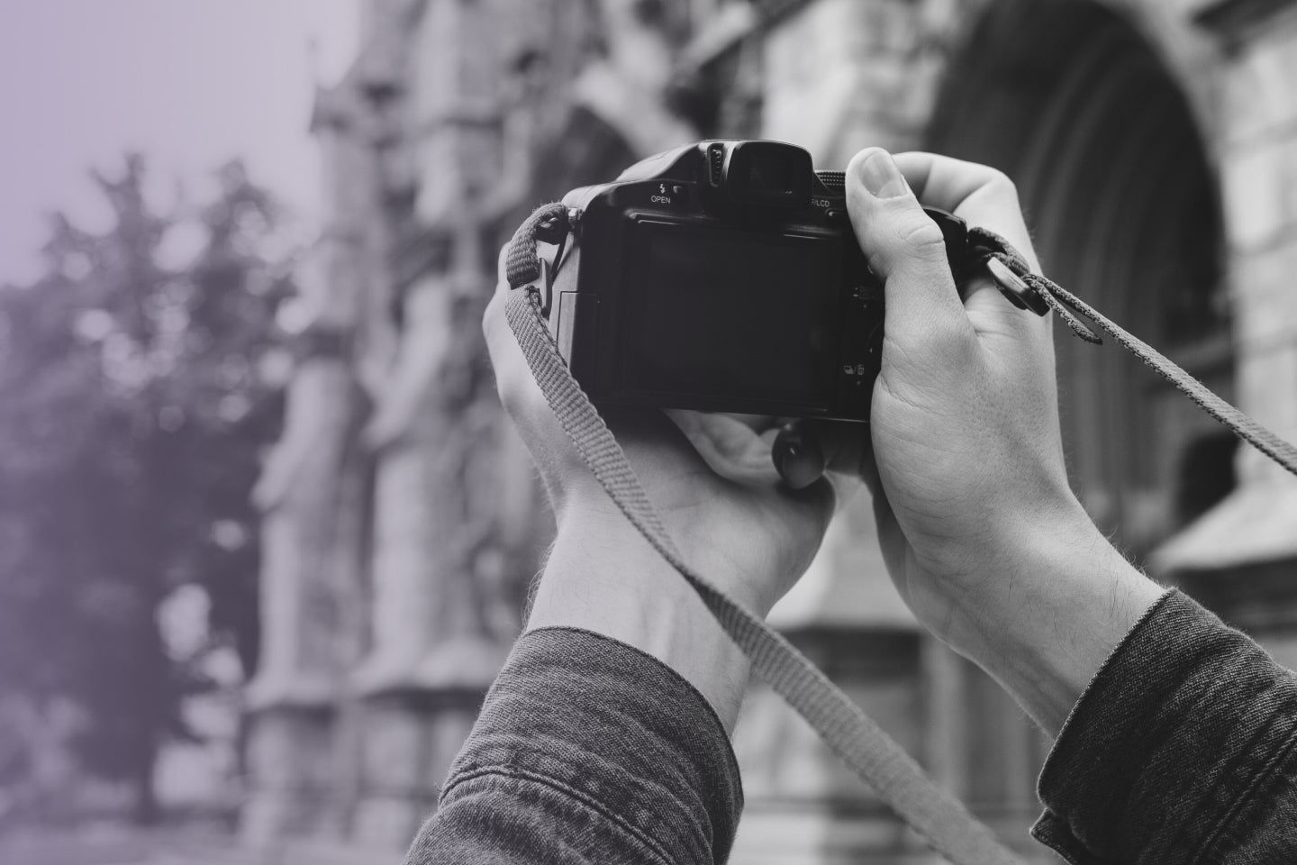 Graues Bildmotiv einer Person die eine Digitalkamera in Richtung eines Gebäudes hält, um dies zu fotografieren.